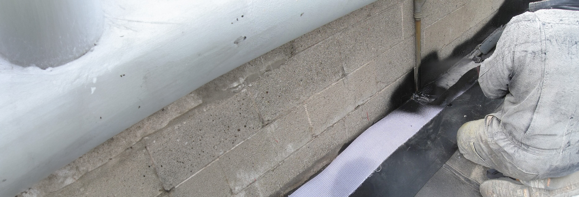 A person is applying a black spray coating containment system to a wall near a pipe, with a textured surface and a white fabric strip visible.