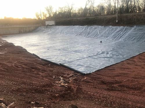 A large, open basin having a containment liner system installed, surrounded by bare soil and trees in the background.